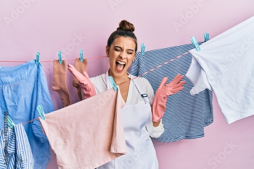 Beautiful brunette young woman washing clothes at clothesline celebrating mad and crazy for success with arms raised and closed eyes screaming excited. winner concept