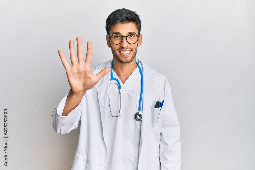 Young handsome man wearing doctor uniform and stethoscope showing and pointing up with fingers number five while smiling confident and happy. Young handsome man wearing doctor uniform and stethoscope showing and pointing up with fingers number five while smiling confident and happy.