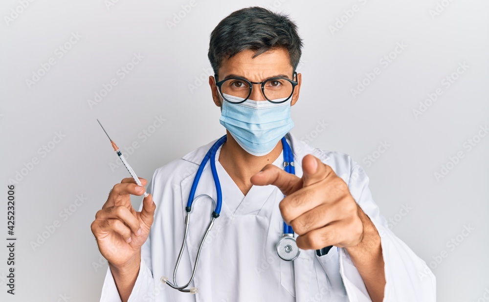 Young handsome man wearing doctor uniform and medical mask holding syringe pointing with finger to the camera and to you, confident gesture looking serious Young handsome man wearing doctor uniform and medical mask holding syringe pointing with finger to the camera and to you, confident gesture looking serious
