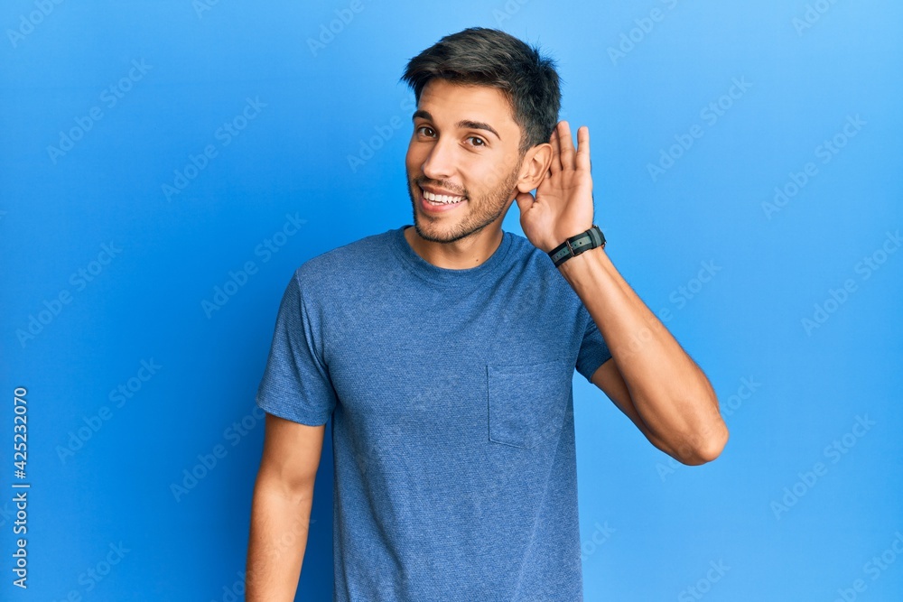 Young handsome man wearing casual tshirt over blue background smiling with hand over ear listening and hearing to rumor or gossip. deafness concept. Young handsome man wearing casual tshirt over blue background smiling with hand over ear listening and hearing to rumor or gossip. deafness concept.