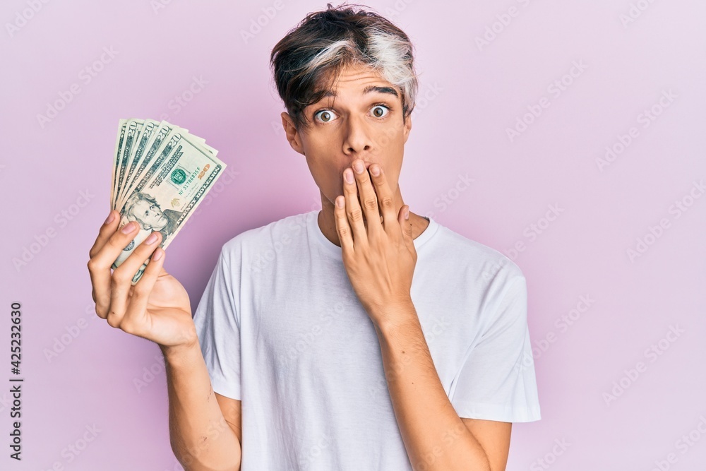 Young hispanic man holding 20 dollars banknote covering mouth with hand, shocked and afraid for mistake. surprised expression Young hispanic man holding 20 dollars banknote covering mouth with hand, shocked and afraid for mistake. surprised expression