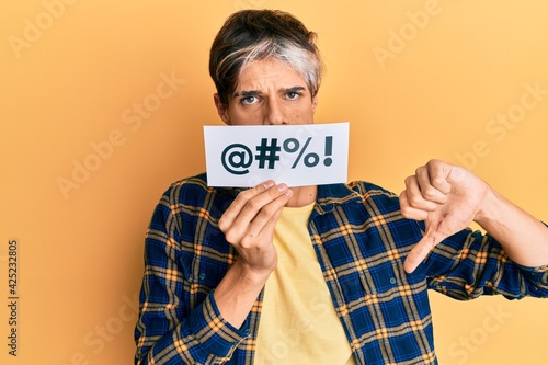 Young hispanic man covering mouth with insult message paper with angry face, negative sign showing dislike with thumbs down, rejection concept