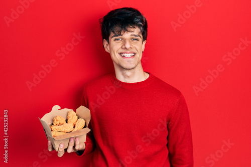 Handsome hipster young man eating chicken wings looking positive and happy standing and smiling with a confident smile showing teeth