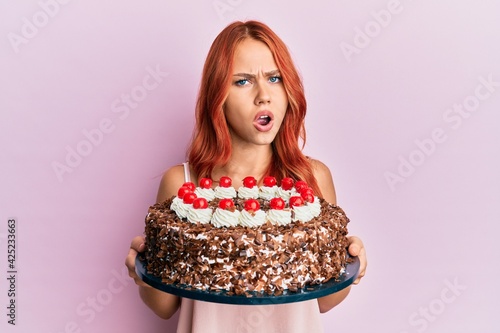 Young redhead woman celebrating birthday with cake in shock face, looking skeptical and sarcastic, surprised with open mouth