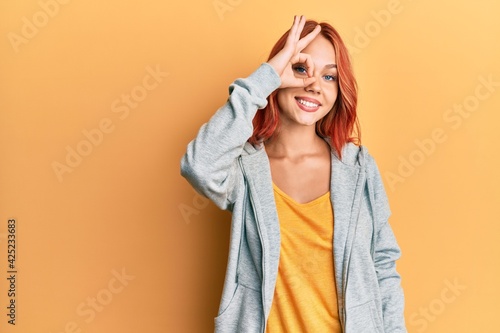 Young beautiful redhead woman wearing casual sporty sweatshirt smiling happy doing ok sign with hand on eye looking through fingers