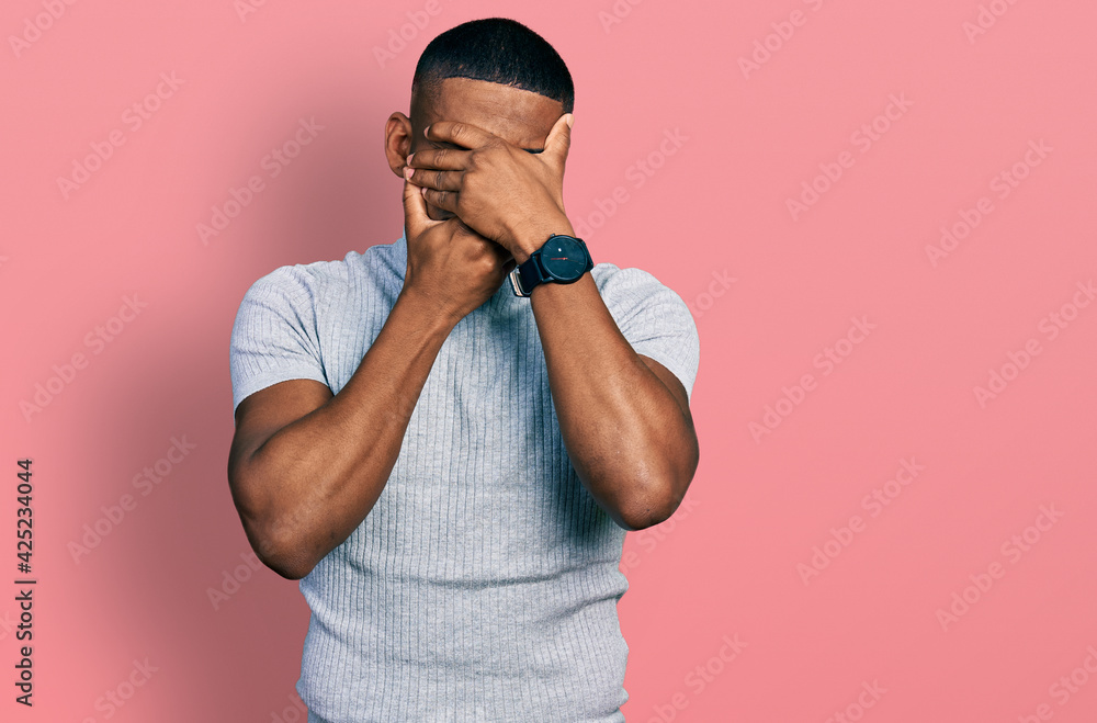 Young black man wearing casual t shirt covering eyes and mouth with hands, surprised and shocked. hiding emotion Young black man wearing casual t shirt covering eyes and mouth with hands, surprised and shocked. hiding emotion
