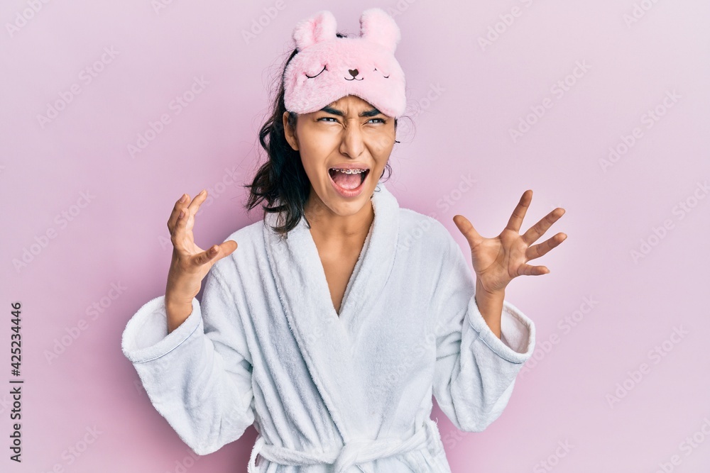 Hispanic teenager girl with dental braces wearing sleep mask and robe crazy and mad shouting and yelling with aggressive expression and arms raised. frustration concept. Hispanic teenager girl with dental braces wearing sleep mask and robe crazy and mad shouting and yelling with aggressive expression and arms raised. frustration concept.