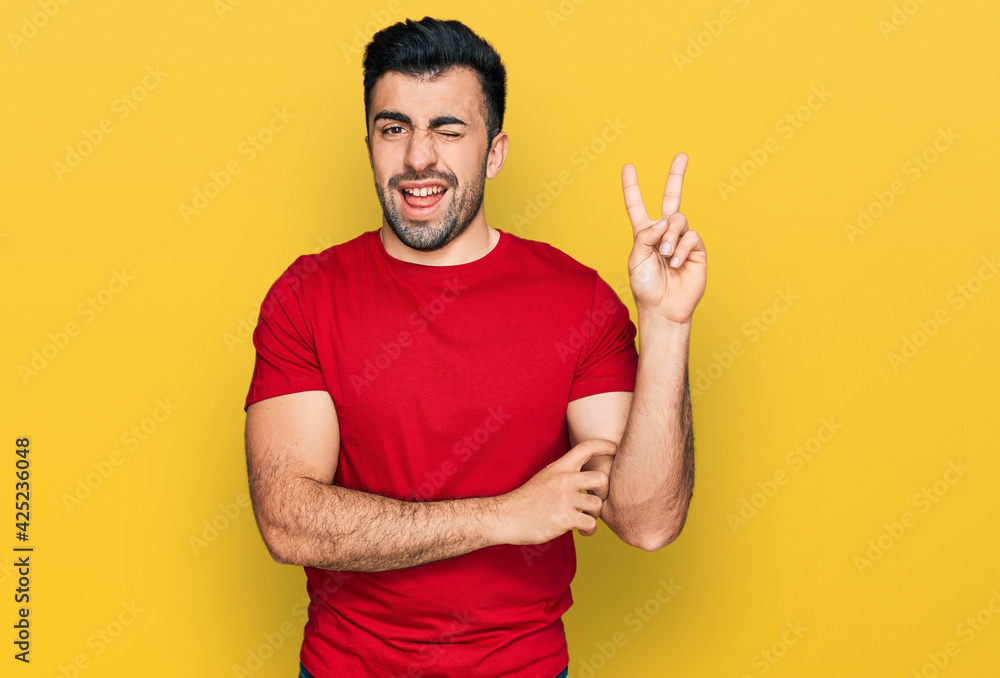 Hispanic man with beard wearing casual red t shirt smiling with happy face winking at the camera doing victory sign. number two. Hispanic man with beard wearing casual red t shirt smiling with happy face winking at the camera doing victory sign. number two.