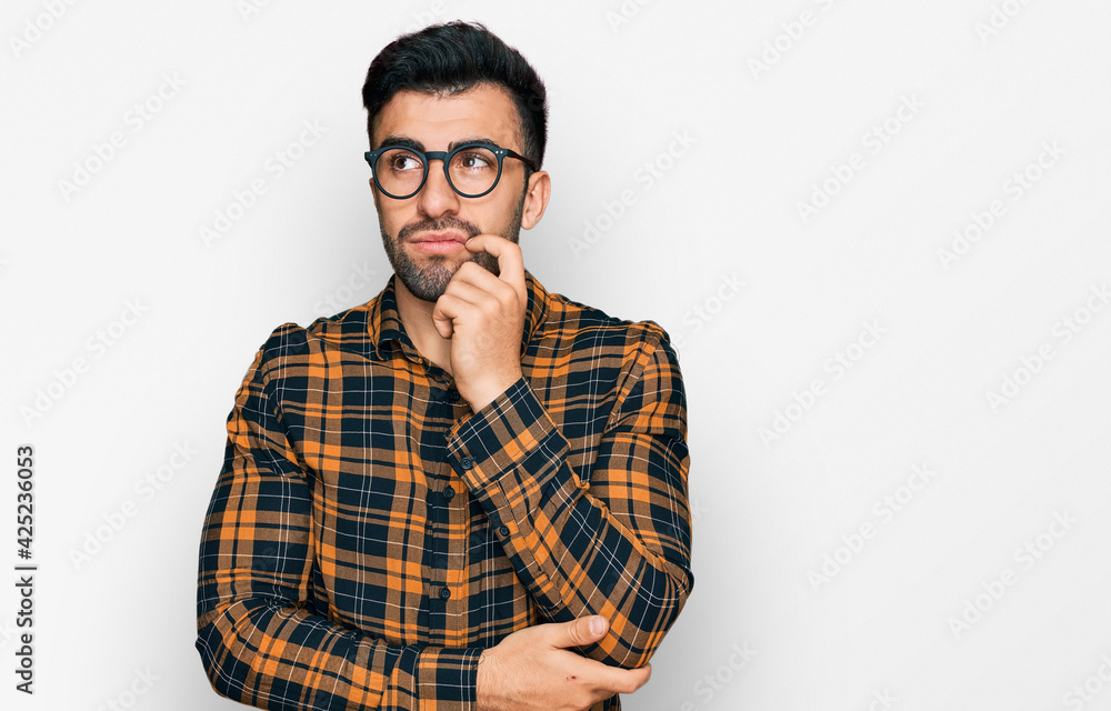 Hispanic man with beard wearing casual clothes and glasses serious face thinking about question with hand on chin, thoughtful about confusing idea Hispanic man with beard wearing casual clothes and glasses serious face thinking about question with hand on chin, thoughtful about confusing idea