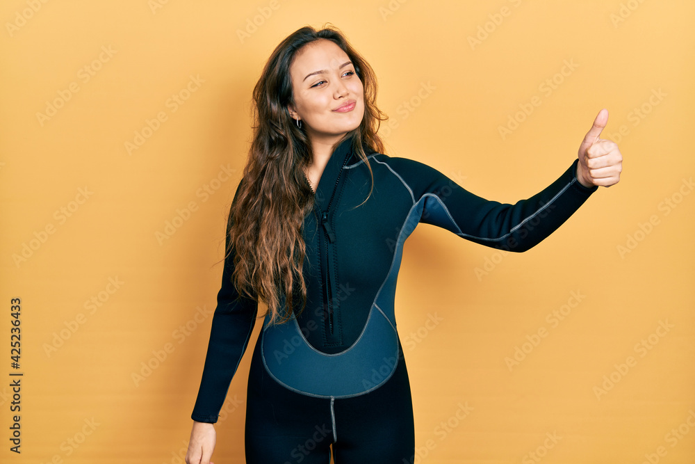 Young hispanic girl wearing diver neoprene uniform looking proud, smiling doing thumbs up gesture to the side Young hispanic girl wearing diver neoprene uniform looking proud, smiling doing thumbs up gesture to the side