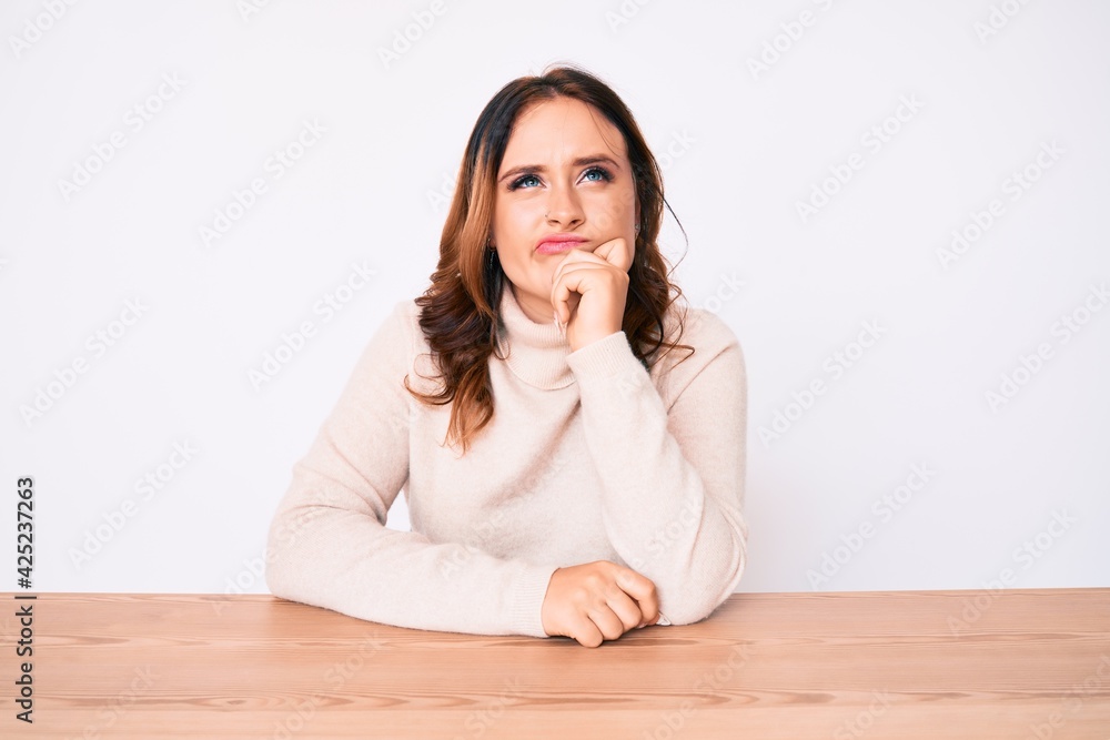 Young beautiful caucasian woman wearing turtleneck sweater sitting on the table thinking concentrated about doubt with finger on chin and looking up wondering Young beautiful caucasian woman wearing turtleneck sweater sitting on the table thinking concentrated about doubt with finger on chin and looking up wondering