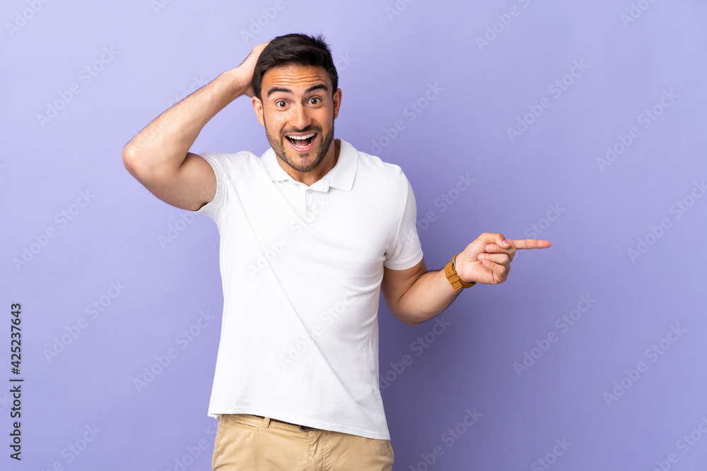 Young handsome man isolated on purple background surprised and pointing finger to the side Young handsome man isolated on purple background surprised and pointing finger to the side