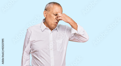 Handsome senior man wearing elegant white shirt smelling something stinky and disgusting, intolerable smell, holding breath with fingers on nose. bad smell © Krakenimages.com