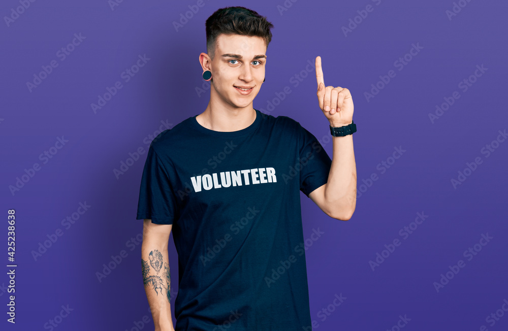 Fototapeta premium Young caucasian boy with ears dilation wearing volunteer t shirt with a big smile on face, pointing with hand finger to the side looking at the camera.