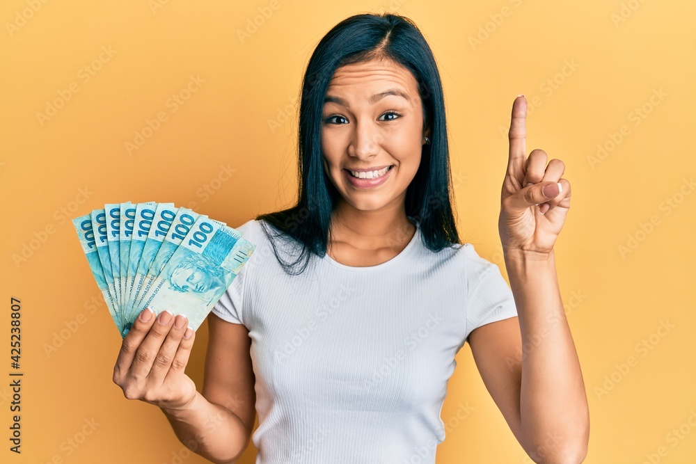 Beautiful hispanic woman holding 100 brazilian real banknotes smiling with an idea or question pointing finger with happy face, number one Beautiful hispanic woman holding 100 brazilian real banknotes smiling with an idea or question pointing finger with happy face, number one