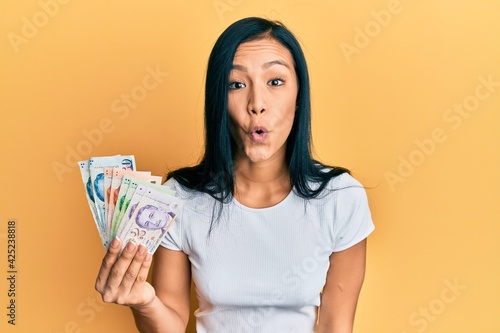 Beautiful hispanic woman holding singapore dollars banknotes scared and amazed with open mouth for surprise, disbelief face