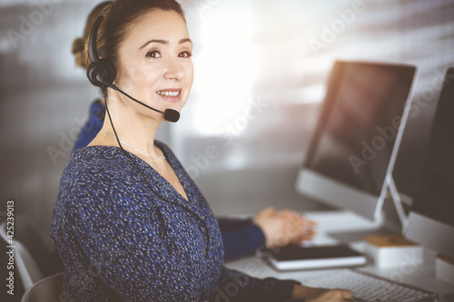 Two busineswomen have conversations with the clients by headsets, while sitting at the desk in a sunny modern office. Diverse people group in a call center. Telemarketing and customer service photo