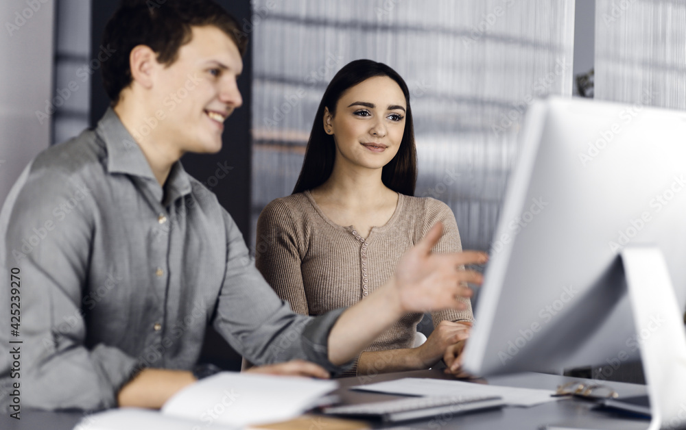 Friendly young businessman and programmer in a green shirt is working on computer, while sitting together at the desk with a female colleague in a modern office. Focus on woman. Concept of successful Friendly young businessman and programmer in a green shirt is working on computer, while sitting together at the desk with a female colleague in a modern office. Focus on woman. Concept of successful