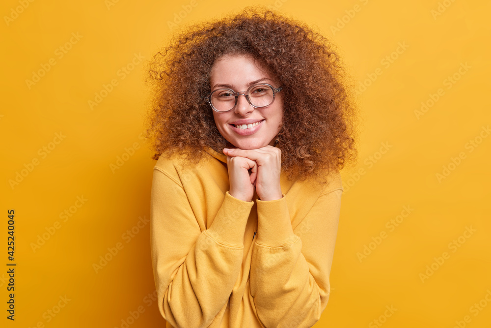 Smiling beautiful teenage girl has natural bushy curly hair smiles toothily keeps hands under chin wears spectacles casual hoodie in one tone with background being pleased by hearing good news Smiling beautiful teenage girl has natural bushy curly hair smiles toothily keeps hands under chin wears spectacles casual hoodie in one tone with background being pleased by hearing good news