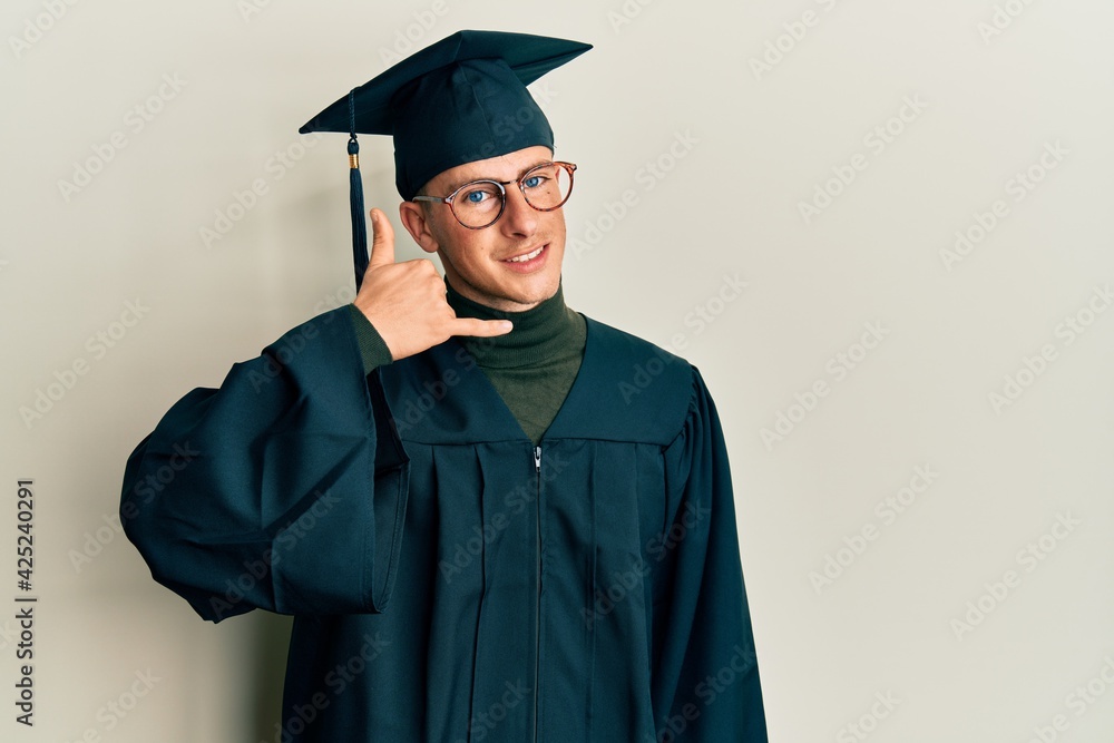 Young caucasian man wearing graduation cap and ceremony robe smiling doing phone gesture with hand and fingers like talking on the telephone. communicating concepts.