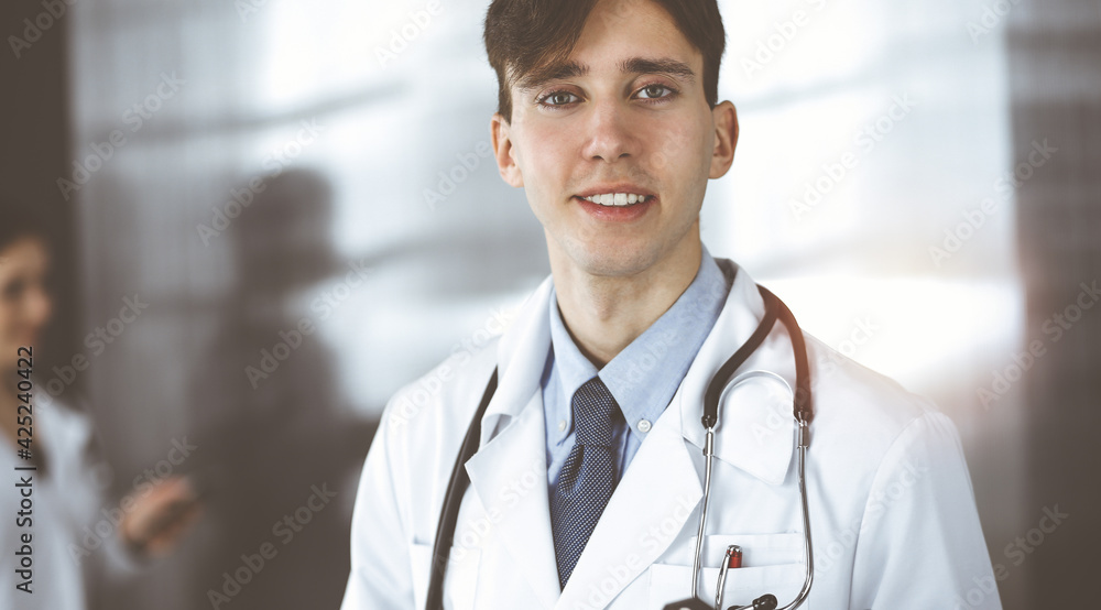 Friendly young male doctor standing with clipboard in modern clinic. Female colleague is busy with filling up of medication records form at the background of physician. Medicine concept Friendly young male doctor standing with clipboard in modern clinic. Female colleague is busy with filling up of medication records form at the background of physician. Medicine concept