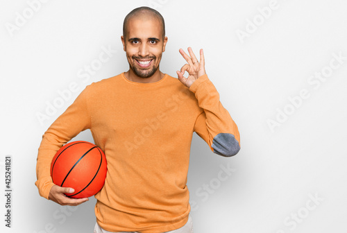 Hispanic adult man holding basketball ball doing ok sign with fingers, smiling friendly gesturing excellent symbol