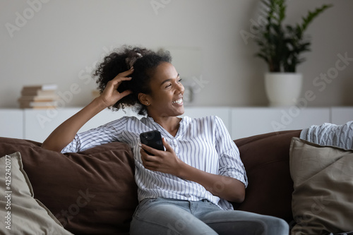 Tableau sur toile Smiling young African American woman sit relax on couch at home use modern smartphone gadget