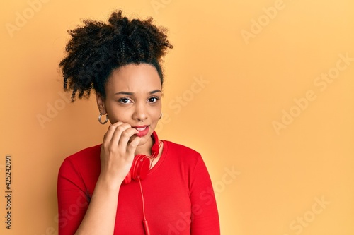 Young african american girl listening to music using headphones looking stressed and nervous with hands on mouth biting nails. anxiety problem.