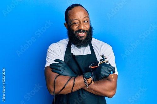 Young african american man tattoo artist wearing professional uniform and gloves holding tattooer machine winking looking at the camera with sexy expression, cheerful and happy face. photo
