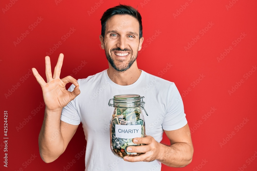 Young handsome man holding charity jar with money doing ok sign with fingers, smiling friendly gesturing excellent symbol Young handsome man holding charity jar with money doing ok sign with fingers, smiling friendly gesturing excellent symbol
