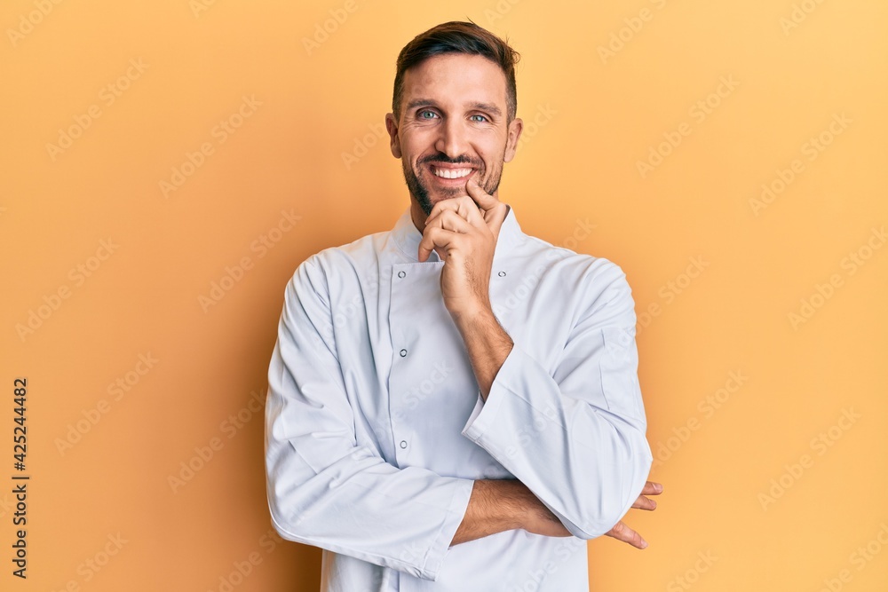 Handsome man with beard wearing professional cook uniform looking confident at the camera with smile with crossed arms and hand raised on chin. thinking positive. Handsome man with beard wearing professional cook uniform looking confident at the camera with smile with crossed arms and hand raised on chin. thinking positive.