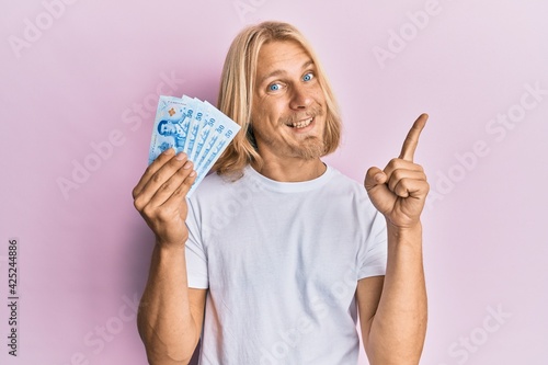 Caucasian young man with long hair holding 50 thai baht banknotes smiling happy pointing with hand and finger to the side