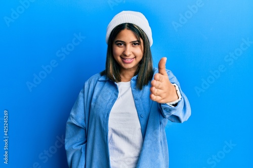 Young latin woman wearing cute wool cap smiling friendly offering handshake as greeting and welcoming. successful business.