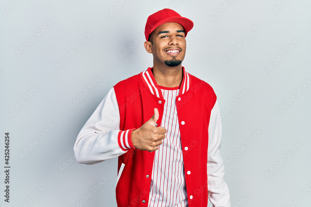 Young african american man wearing baseball uniform doing happy thumbs up gesture with hand. approving expression looking at the camera showing success. Young african american man wearing baseball uniform doing happy thumbs up gesture with hand. approving expression looking at the camera showing success.