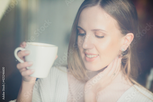 Beautiful young woman is drinking coffee. Attractive woman in a cozy cafe or at home. Pretty woman with a cup of cappuccino in her hands. Close-up portrait. Soft focus and shallow depth of field.