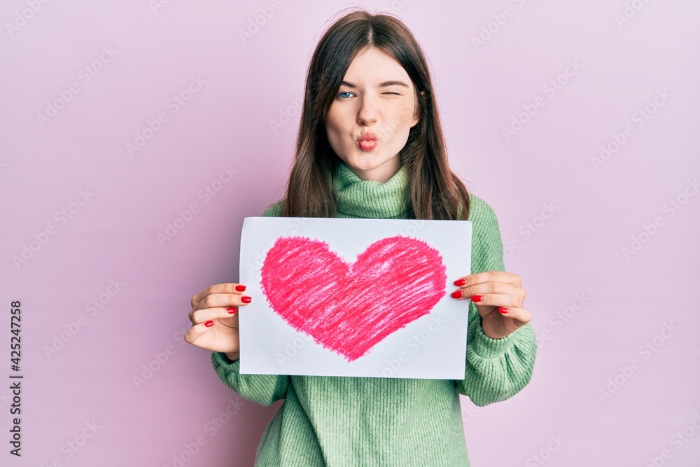 Young beautiful caucasian girl holding heart draw looking at the camera blowing a kiss being lovely and sexy. love expression. Young beautiful caucasian girl holding heart draw looking at the camera blowing a kiss being lovely and sexy. love expression.