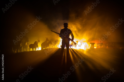 Silhouette of a man (hunter) with rifle standing against group of animals in colorful dark backlight. Decorated with miniatures. Selective focus photo