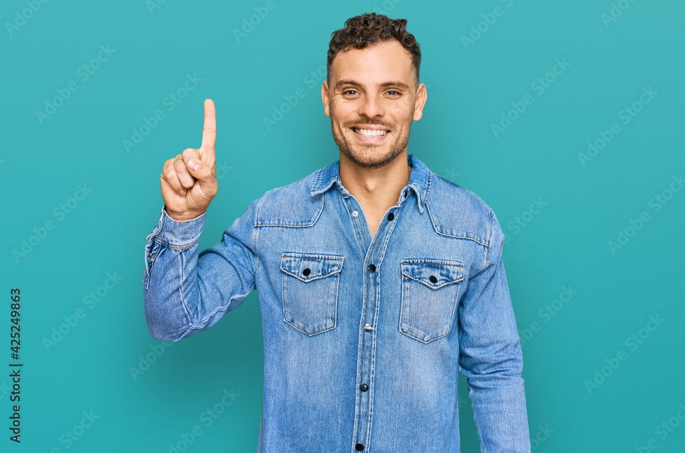 Young hispanic man wearing casual denim jacket showing and pointing up with finger number one while smiling confident and happy. Young hispanic man wearing casual denim jacket showing and pointing up with finger number one while smiling confident and happy.