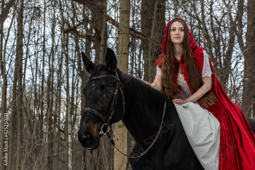 beautiful girl in a white dress and a red cloak with a black horse in the spring forest