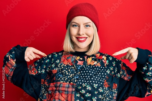 Young blonde woman wearing wool winter cap looking confident with smile on face, pointing oneself with fingers proud and happy.