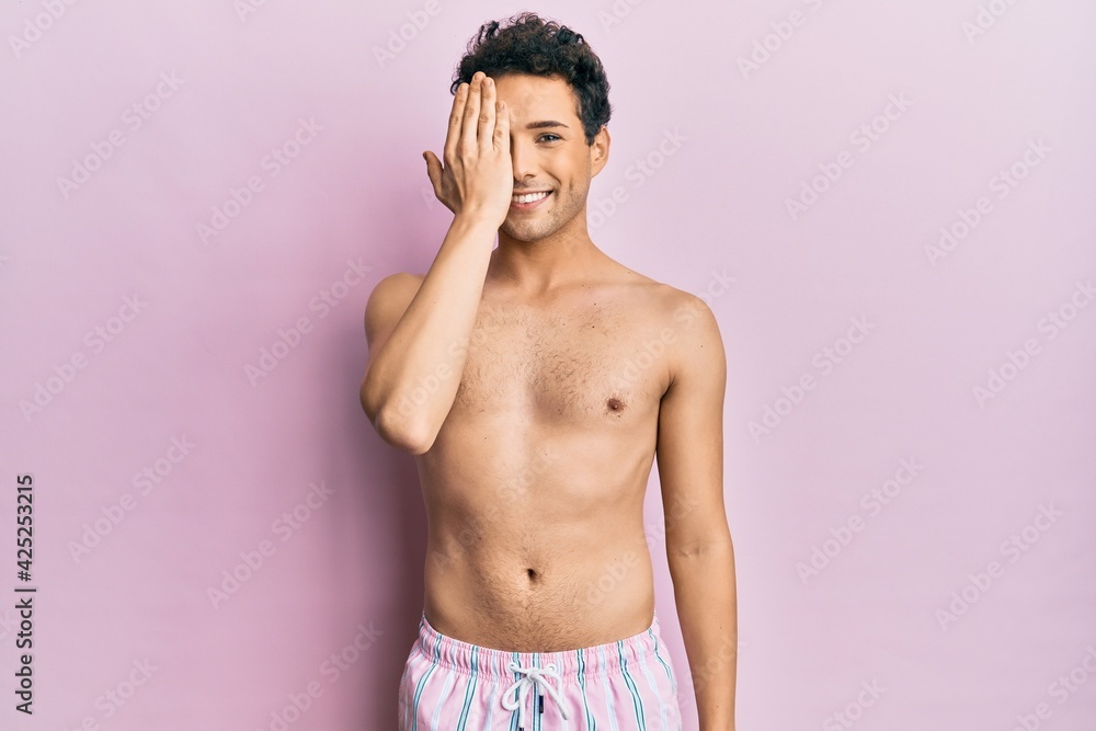 Fototapeta premium Young handsome man wearing swimwear shirtless covering one eye with hand, confident smile on face and surprise emotion.