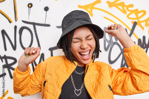 Bilde på lerret Overjoyed teenager in urban outfit raises arms has fun enjoys spare time poses against colorful graffiti wall belongs to youth subculture moves carefree exclaims joyfully