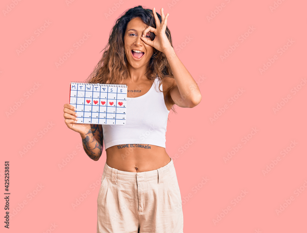 Young hispanic woman with tattoo holding heart calendar smiling happy doing ok sign with hand on eye looking through fingers Young hispanic woman with tattoo holding heart calendar smiling happy doing ok sign with hand on eye looking through fingers