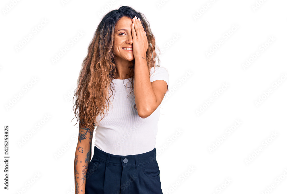 Young hispanic woman with tattoo wearing casual white tshirt covering one eye with hand, confident smile on face and surprise emotion. Young hispanic woman with tattoo wearing casual white tshirt covering one eye with hand, confident smile on face and surprise emotion.