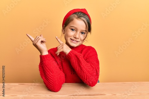 Little beautiful girl wearing casual clothes sitting on the table smiling and looking at the camera pointing with two hands and fingers to the side.