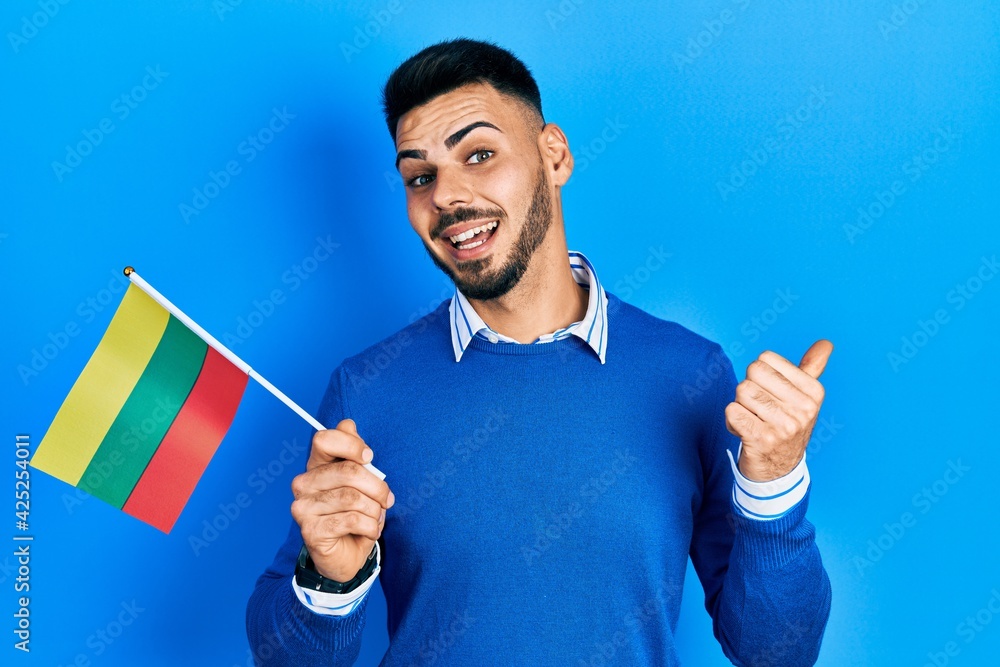Young hispanic man with beard holding lithuania flag pointing thumb up to the side smiling happy with open mouth Young hispanic man with beard holding lithuania flag pointing thumb up to the side smiling happy with open mouth