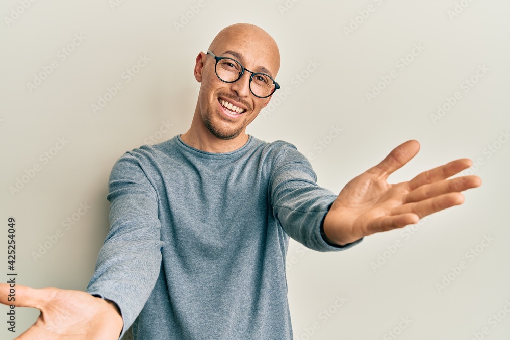 Bald man with beard wearing casual clothes and glasses looking at the camera smiling with open arms for hug. cheerful expression embracing happiness. Bald man with beard wearing casual clothes and glasses looking at the camera smiling with open arms for hug. cheerful expression embracing happiness.