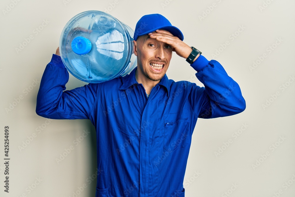 Bald courier man with beard holding a gallon bottle of water for delivery stressed and frustrated with hand on head, surprised and angry face