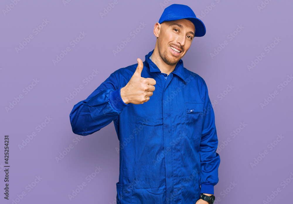 Bald man with beard wearing builder jumpsuit uniform doing happy thumbs up gesture with hand. approving expression looking at the camera showing success.