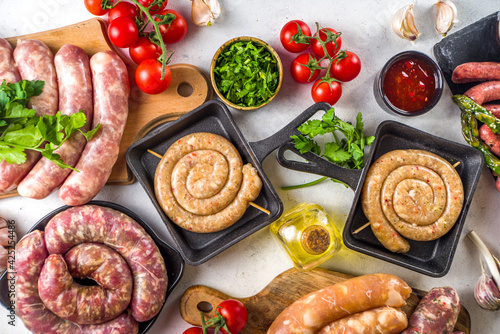 Set of different raw beef  pork and chicken meat sausages on cooking table with spices  sauces and vegetables for grilling. Bbq party concept  white background top view copy space